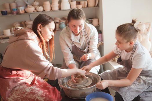 Professional potter teaching young woman with child throwing pot on pottery wheel