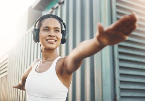 One fit young hispanic woman stretching arms in warrior pose for warmup to prevent injury while exercising in an urban setting outdoors. Happy and motivated female athlete listening to music with headphones while preparing body and mind for training workout or run.