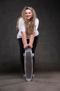 Beautiful fat woman with a skateboard on a gray background looks at the camera and smiles.