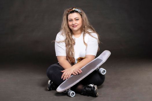 Beautiful fat woman with a skateboard on a gray background looks at the camera and smiles.