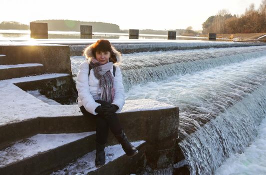 A woman sitting in front of a waterfall. Portrait of a sitting pretty elderly woman in a white jacket with scarf and black gloves in front of a waterfall at a reservoir looking at the camera.