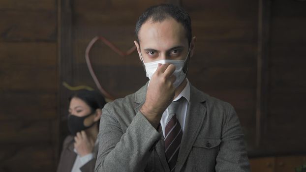 Middle Eastern Man In Suit And Tie Adjusts Protective Mask With Hand, Serious Businessman Looking At Camera While Standing In Modern Office, Office Workers In Medical Masks Working Despite Quarantine