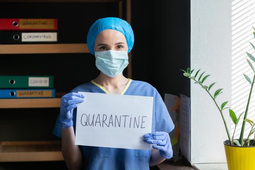 Young Woman Medical worker In Blue Protective Uniform declares quarantine Showing This Word Written By Marker On Paper, Cheerful Woman In Mask Looks At Camera Standing On Medical Office Background