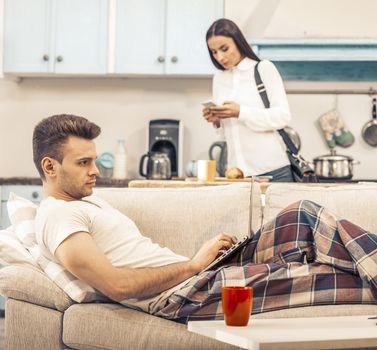 Freelancer Man Staying And Working From Home, Caucasian Man Works With Laptop Lying On The Couch While His Woman In White Blouse Going To Office Work, Toned Image