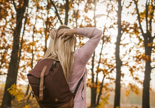 Young Woman With A Backpack Turned His Face To The Sun, Admires The Autumn Forest, Rear View Of Caucasian Blonde Putting Her Hands On Back Of Her Head While Relaxing On Autumn Trip