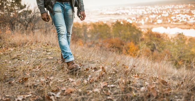 Man Hikes On A Hill, Young Man In Jeans With Backpack Is Walking Along The Autumn Grass With Beauty Landscape In Background, Close Up Of Men's Legs In Blue Jeans And Leather Boots.