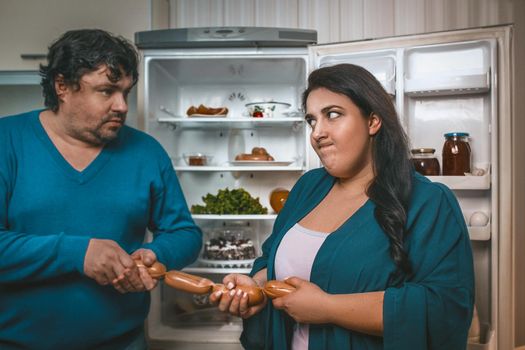 Plus Size Man And Woman Take Away Sausages From Each Other, Hungry Caucasian Body Positive Couple Fight For Food Near Open Refrigerator In Kitchen, Selective Focus On Brunette Woman