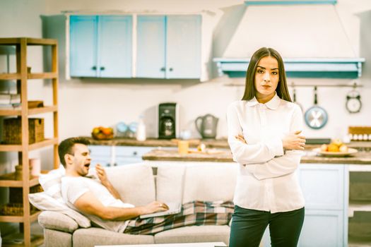 Bored Couple At Home Awaiting Quarantine Ending, Selective Focus On Sad Woman Standing In Foreground And Looking At Camera, Her Husband Yawns Lying On Sofa, Toned Image