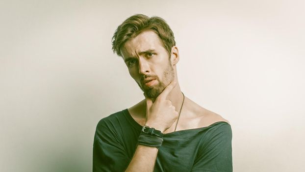 Handsome Man Wrinkled His Forehead Thoughtfully Touching His Chin With His Hand, Bearded Caucasian Man In Black Casual Clothing Poses On A Gray Background