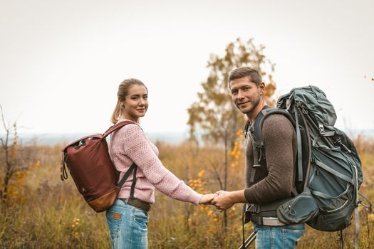 Young Couple Of Travelers Smiles Holding Hands Outdoors, Happy Backpacker Man And Woman Smiles Looking Back At Camera Standing Against The Background Of Autumn Field And Sky