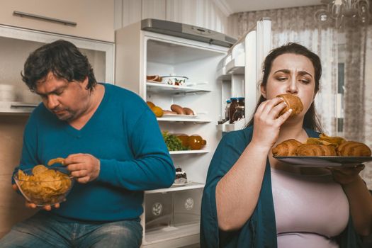 Overweight Family Overeating Unhealthy Food In Kitchen, Man And Woman Standing Near Open Frig Holding Bowl With Potato Chips And Plate Of Croissants While Overeating At Bedtime