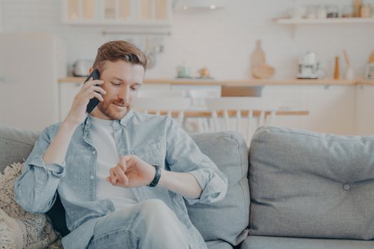 Young man talking on phone while checking time on his wristwatch, sitting on couch in his apartment with crossed legs, agreeing on meeting with friend, choosing best time to meet