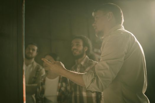 Diverse Group Of Students Or Entrepreneurs Studying Map While Standing In Backlight, Focus On Hand Of Caucasian Man In Foreground Pointing With Index Finger At Map Or Poster Hanging On Office Wall