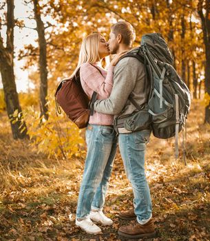 Kiss In Autumn Forest Outdoors, Young Loving Couple With Large Backpacks Kisses On Warm Autumn Day Against The Background Of Golden Forest In Sunlight, Full Length Profile View