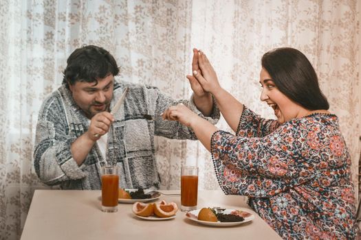 Body Positive Woman Tries To Feed Her Husband Healthy Food, Smiling Woman Gives A Man High Five With One Hand And Holding Fork With Salad In Other Hand, She Persuades Her Husband To Open His Mouth