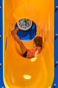 Young girl in side view relaxing inside a yellow plastic pipe or tube on playground climbing frame and slide