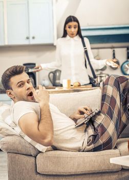 Bored Man In Home Clothes Yawns While Lying On Sofa With Computer, His Wife Makes Helpless Gesture On Blurred Background, Focus On Caucasian Man In Foreground, He Need To Stay At Home, Toned Image
