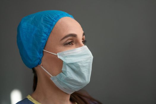 Female Medic In Protective Mask And Blue Uniform Looks At Right Side With Copy Space, Profile View Of Nurse Or Doctor Turning Her Face To Light, Portrait Of Young Caucasian Woman On Gray Background