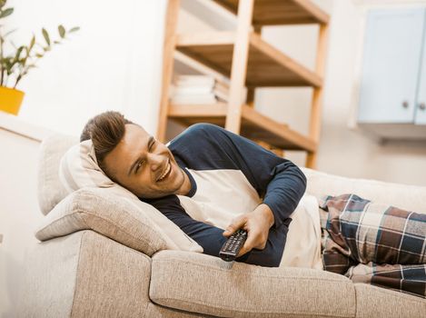 Happy Man Relaxing At Home, He Watching TV Lying On Sofa, Smiling Man In Home Clothes Winks Joyfully While Staying At Home During Quarantine, Toned Image