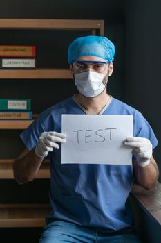 Male Nurse In Protective Medical Uniform Suggests Making A Test For Coronavirus Infection, Serious Middle Eastern Man In Ptotective Wear Demonstrates A Slogan Written On White Paper