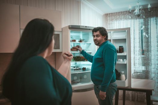 Angry Wife Forbids Her Husband To Take Food, Body Positive Man Stands Near Opened Refrigerator Grabbing Sausages With His Hand, Rear View Of His Wife Showing Him A Fist Standing In Foreground