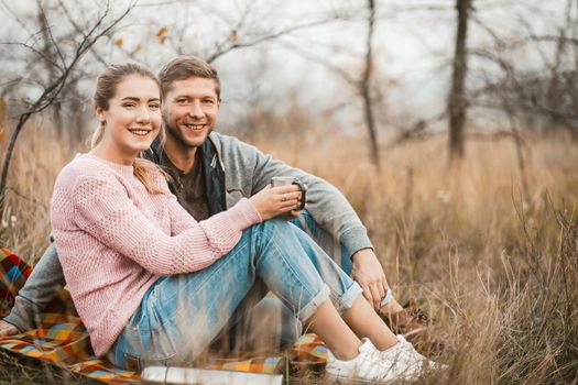 Smiling Couple Drink Coffee Or Tea Outdoors, Cheerful Man And Woman Looks At Camera Holding Cups With Hot Drink While Sitting On Grass, Picnic In Nature Concept, Side View