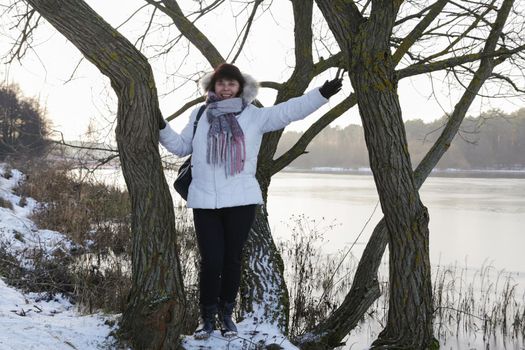 European woman in white winter jacket with scarf standing near tree in front of river.