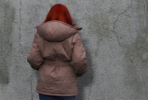 A plus-size girl with red hair in a brown jacket against an wall with her hands in her pockets from the back.