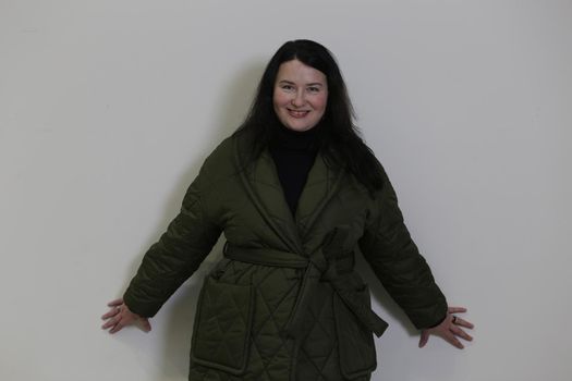 Cheerful brunette plus size in a green winter coat stands leaning against a white wall.