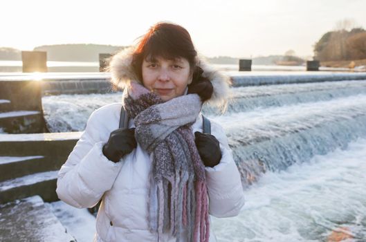 Portrait of a woman in a white jacket with scarf and black gloves against the background of a river and waterfall. Woman against the background of a water landscape looking at the camera.