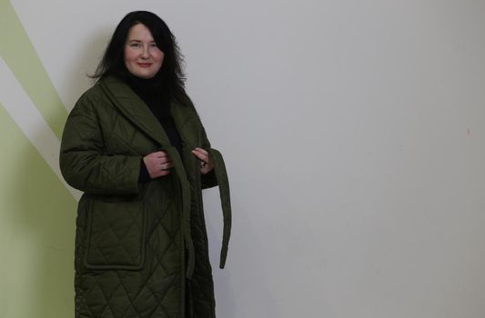 .A pretty plus size woman with black hair stands in a green raincoat against a white wall. A woman in an unbuttoned coat.