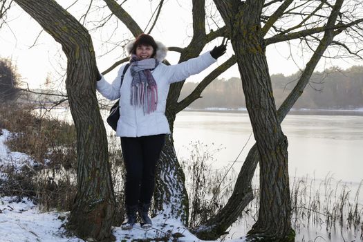 Pretty woman in white jacket with scarf standing near tree in winter river background.