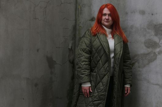 A plus-size girl with red hair in a green jacket poses in front of an old wall with damaged plaster.