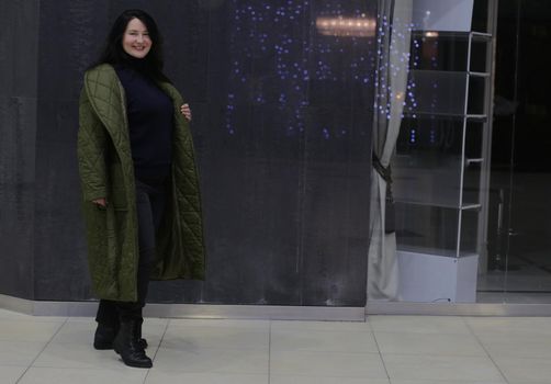 Full length image of a woman in a shop interior. A beautiful plus size woman in a black sweater, trousers and a green coat smiles and looks at the camera.