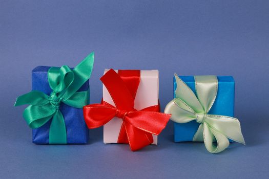 Two blue and one white paper gift boxes with ribbons on blue background. Three beautiful boxes wrapped in colored paper with bows.