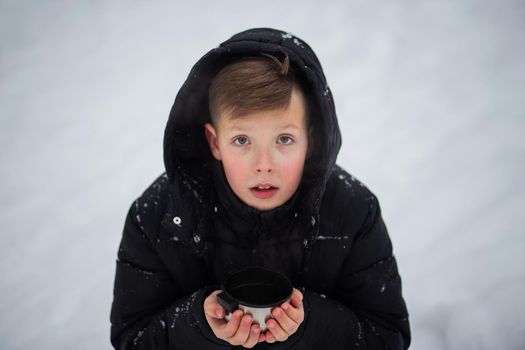 A cute teenage boy in winter clothes with a hood on his head is holding a metal mug in his hands. A handsome boy with a mug in his hands looks at the camera against a background of snow.