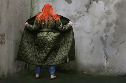 A plus-size girl with red hair standing in an unbuttoned green coat in front of an old wall with peeling plaster, back view...Back of a girl in an open long raincoat facing a full-length wall.