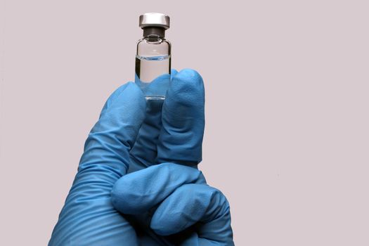 A hand in a blue medical glove close-up holding a vaccine against a light background. The concept of the fight against Covid19..