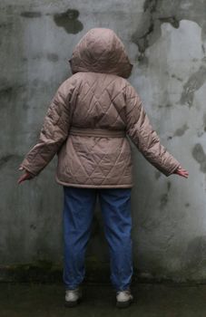 Brown haired plus size girl in brown jacket in blue jeans against old concrete wall back view.
