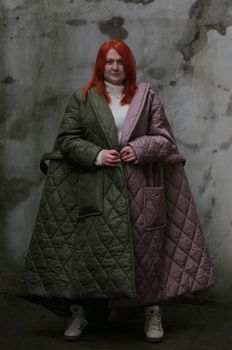 Full-length portrait of a redheaded plus-size girl wearing green and brown long coats and looking at the camera.