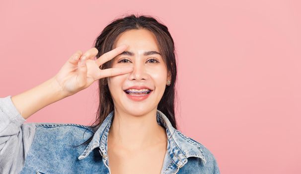 Asian happy portrait beautiful cute young woman wear denim smile standing showing finger making v-sign victory symbol near eye looking to camera studio shot isolated on pink background with copy space