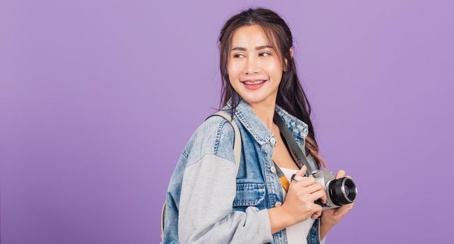 Attractive portrait happy Asian beautiful young woman smiling excited wear denims and bag holding vintage photo camera, female traveler female photographer, studio shot isolated on purple background