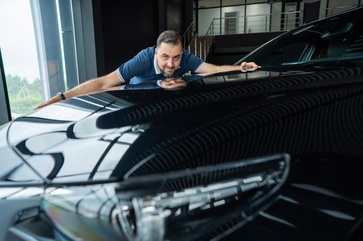 Happy caucasian man hugging the hood of his new car in a car dealership