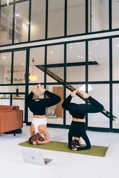 Two beautiful young woman working out indoors, doing yoga exercise on mat, supported headstand, salamba sirsasana, rear view, full length