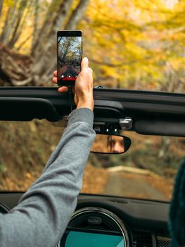 Man takes a vertical video on a mobile phone from the car on the road in the autumn beautiful forest.