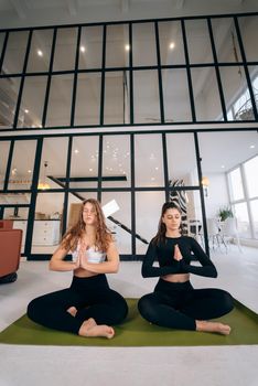 Two young women meditating in lotus pose with hands in namaste. Yoga