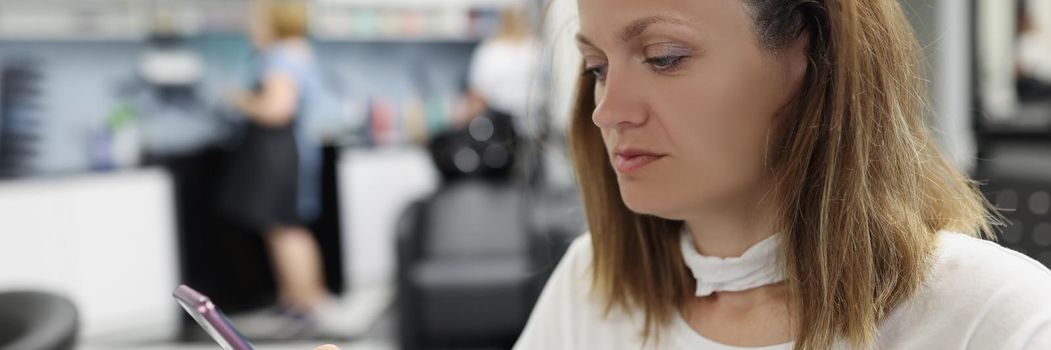 Portrait of woman client sit at hairdresser, woman use smartphone while waiting for hair to dye. Female enjoy beauty procedure. Wellness, self care concept