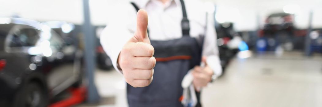Close-up of male worker in uniform showing thumbs up posing in garage. Fixed car after maintenance in service. Pit stop, service station, handyman concept
