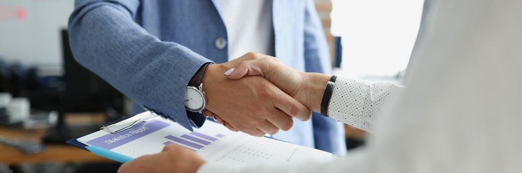 Close-up of partners shake hands over business statistics report paper. Biz partners perform friendly gesture after successful agreement. Contract concept