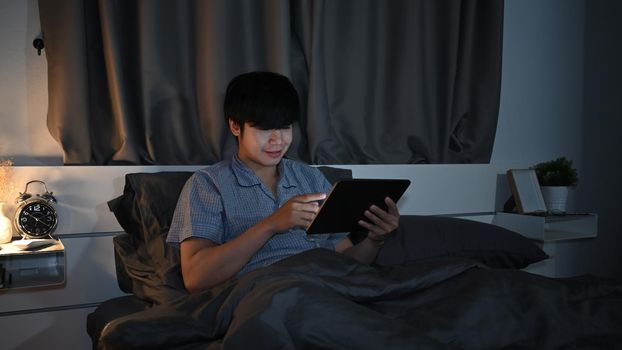 Asian man sitting in bedroom and browsing internet with digital tablet.